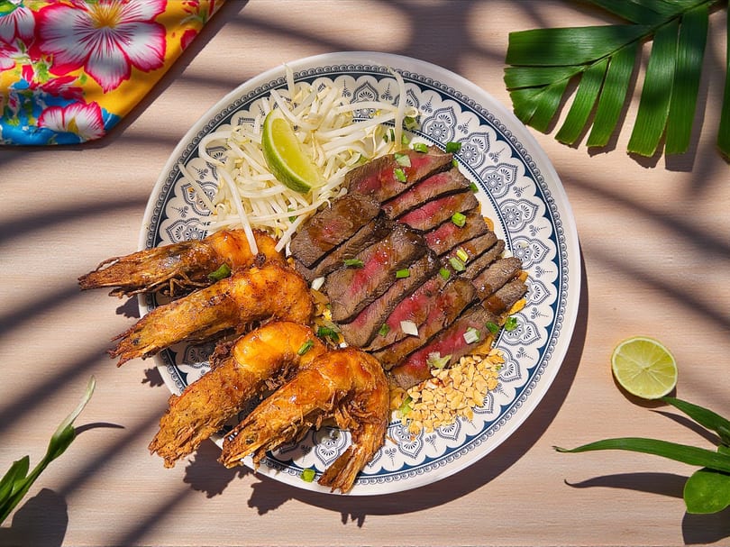 Sliced steak, large prawns, bean sprouts, peanuts, and lime on a decorative plate.