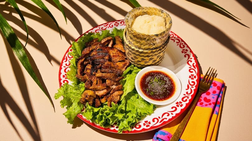 Grilled meat with lettuce, sticky rice in a bamboo basket, and dipping sauce on a decorative plate.