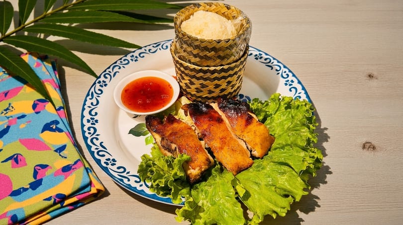 Grilled chicken with sticky rice in a bamboo basket, sweet chili sauce, and lettuce on a patterned plate.