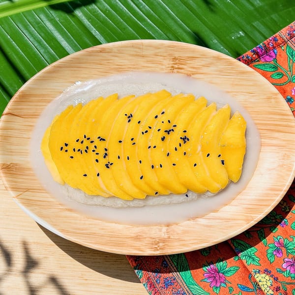 Bright mango sticky rice with black sesame seeds on a wooden plate, banana leaf, and floral napkin.