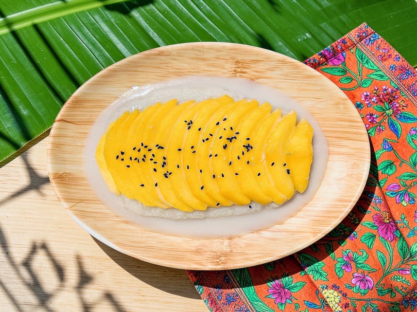 Bright mango sticky rice with black sesame seeds on a wooden plate, banana leaf, and floral napkin.