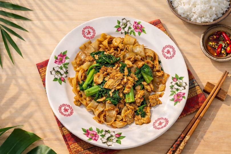 Flat noodles with chicken, green vegetables, rice, and chili sauce on a floral plate.