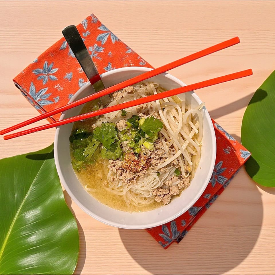 White bowl of noodle soup with ground pork, bean sprouts, and cilantro, with red chopsticks on wood.