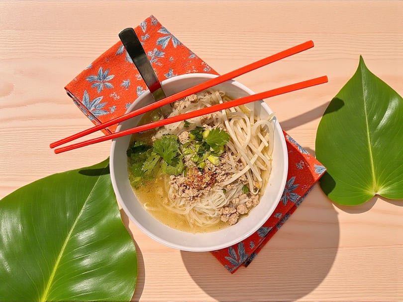 White bowl of noodle soup with ground pork, bean sprouts, and cilantro, with red chopsticks on wood.